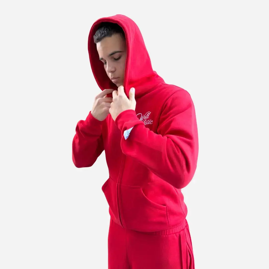 Side view of a model wearing the CELESTIC Red Basic Tracksuit with the hood up. Highlighting the premium heavyweight red fleece zip-up hoodie with white script logo embroidery on the chest and a structured double-lined hood, paired with matching red sweatpants.