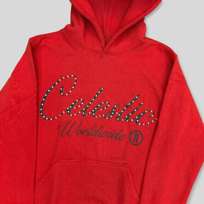 Detailed close-up photograph of the CELESTIC Red Gemstone Hoodie. The focus is on the chest, showcasing the vibrant red premium cotton fleece and the large black screen-printed 'Celestic' script logo, heavily detailed with sparkling rhinestones. The printed text 'Worldwide ®' is clearly visible below the script. The base of the structured hood and the top of the kangaroo pocket frame the logo.