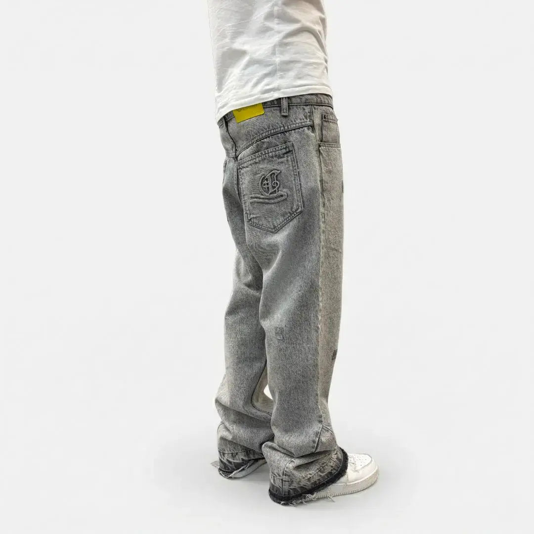 Grey flared jeans back angle view on model. Washed distressed denim pants highlighting the custom back pocket embroidery, yellow brand patch, relaxed fit, and frayed raw hems stacking perfectly over white sneakers.