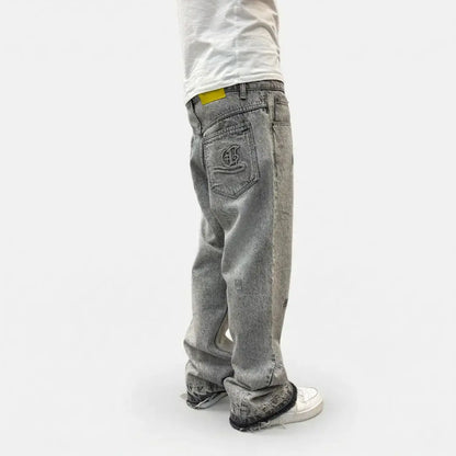Grey flared jeans back angle view on model. Washed distressed denim pants highlighting the custom back pocket embroidery, yellow brand patch, relaxed fit, and frayed raw hems stacking perfectly over white sneakers.