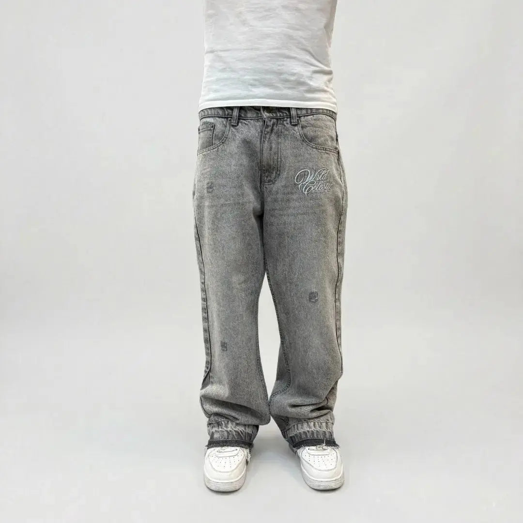 Grey flared jeans front view on model. Washed distressed denim pants featuring a white Celestic embroidered logo on the thigh, subtle rips, lower leg panel stitching, and frayed raw hems styled over white sneakers.