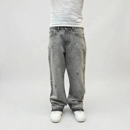 Grey flared jeans front view on model. Washed distressed denim pants featuring a white Celestic embroidered logo on the thigh, subtle rips, lower leg panel stitching, and frayed raw hems styled over white sneakers.