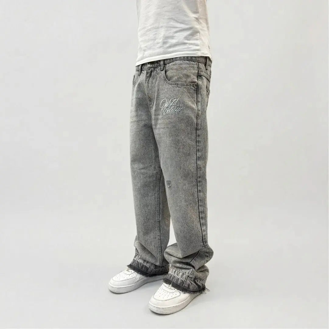 Grey flared jeans side view on model. Washed distressed denim pants highlighting the front thigh embroidery, leg panel stitching, relaxed fit, and frayed raw hems stacking perfectly over white sneakers.