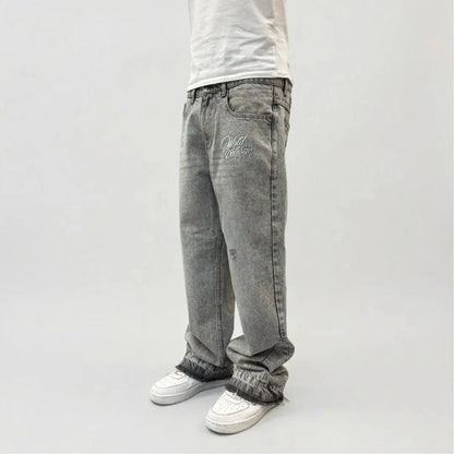 Grey flared jeans side view on model. Washed distressed denim pants highlighting the front thigh embroidery, leg panel stitching, relaxed fit, and frayed raw hems stacking perfectly over white sneakers.