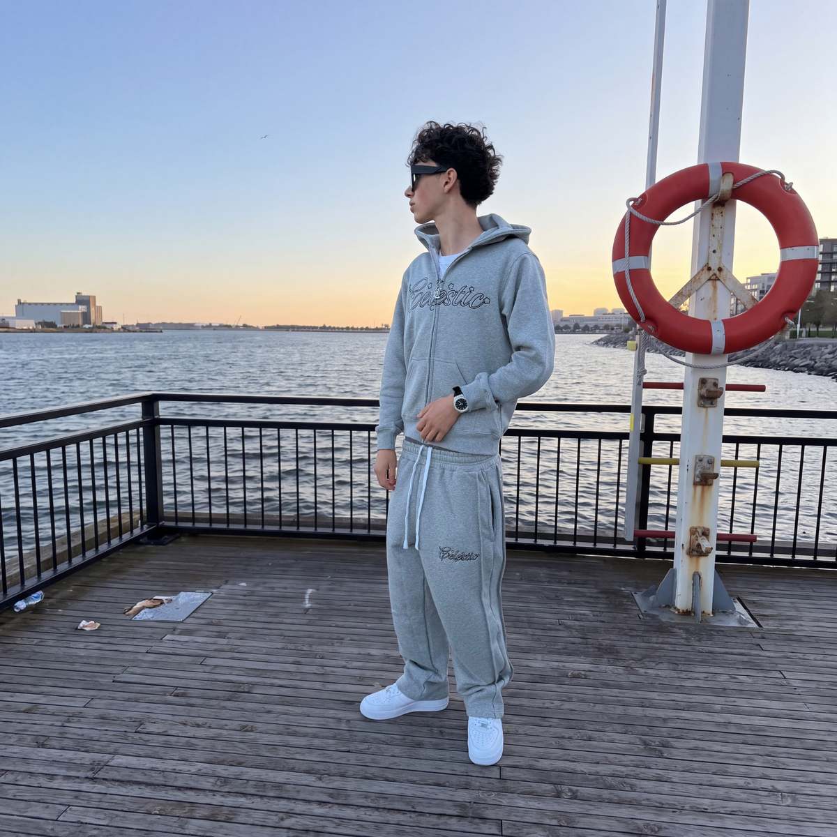 Model wearing the CELESTIC Grey Outline Tracksuit on a pier at sunset, with Celestic outline embroidery on the zip-up hoodie and pants.