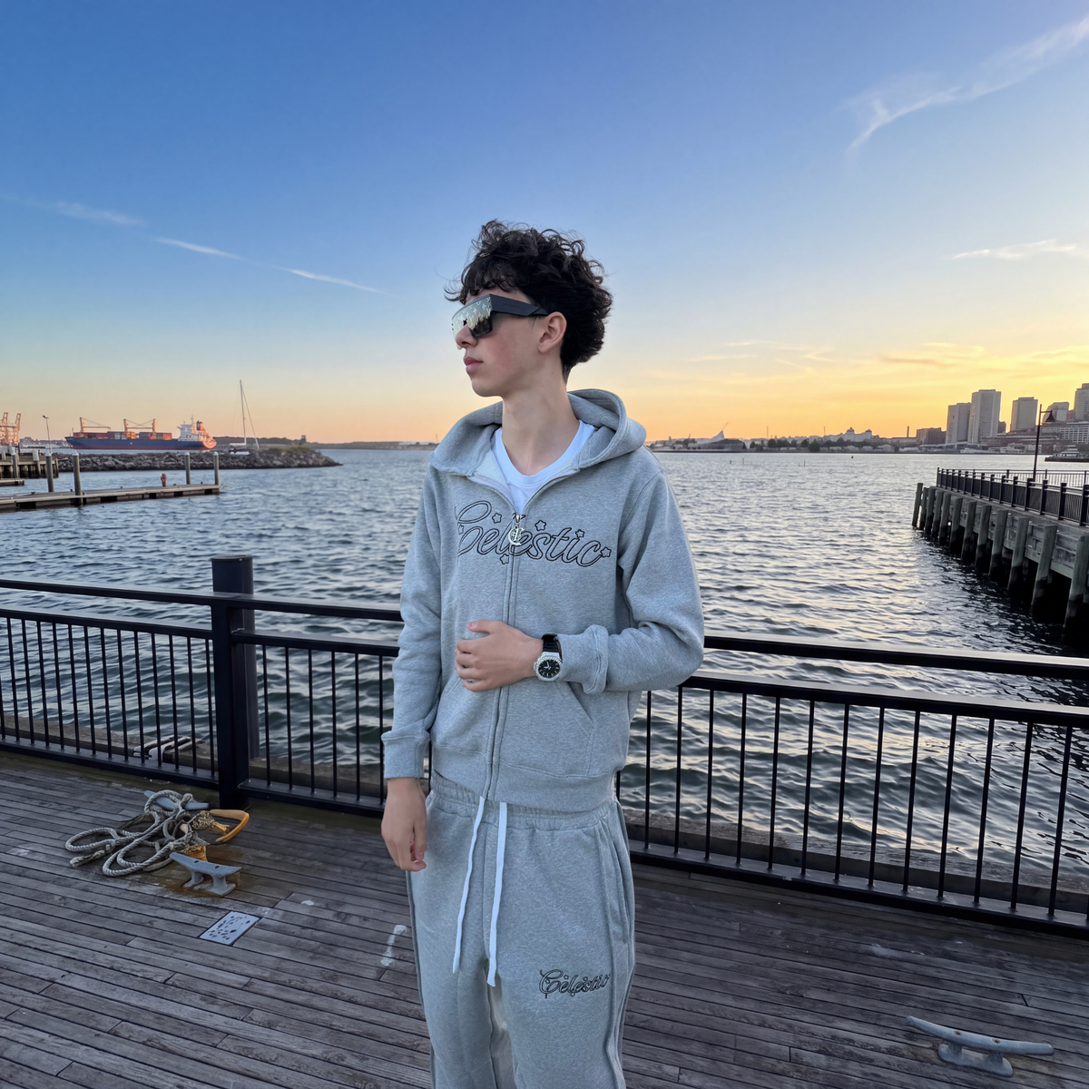 Model in the CELESTIC Grey Outline zip-up hoodie and pants on a pier at sunset, showing the Celestic outline embroidery across the chest.