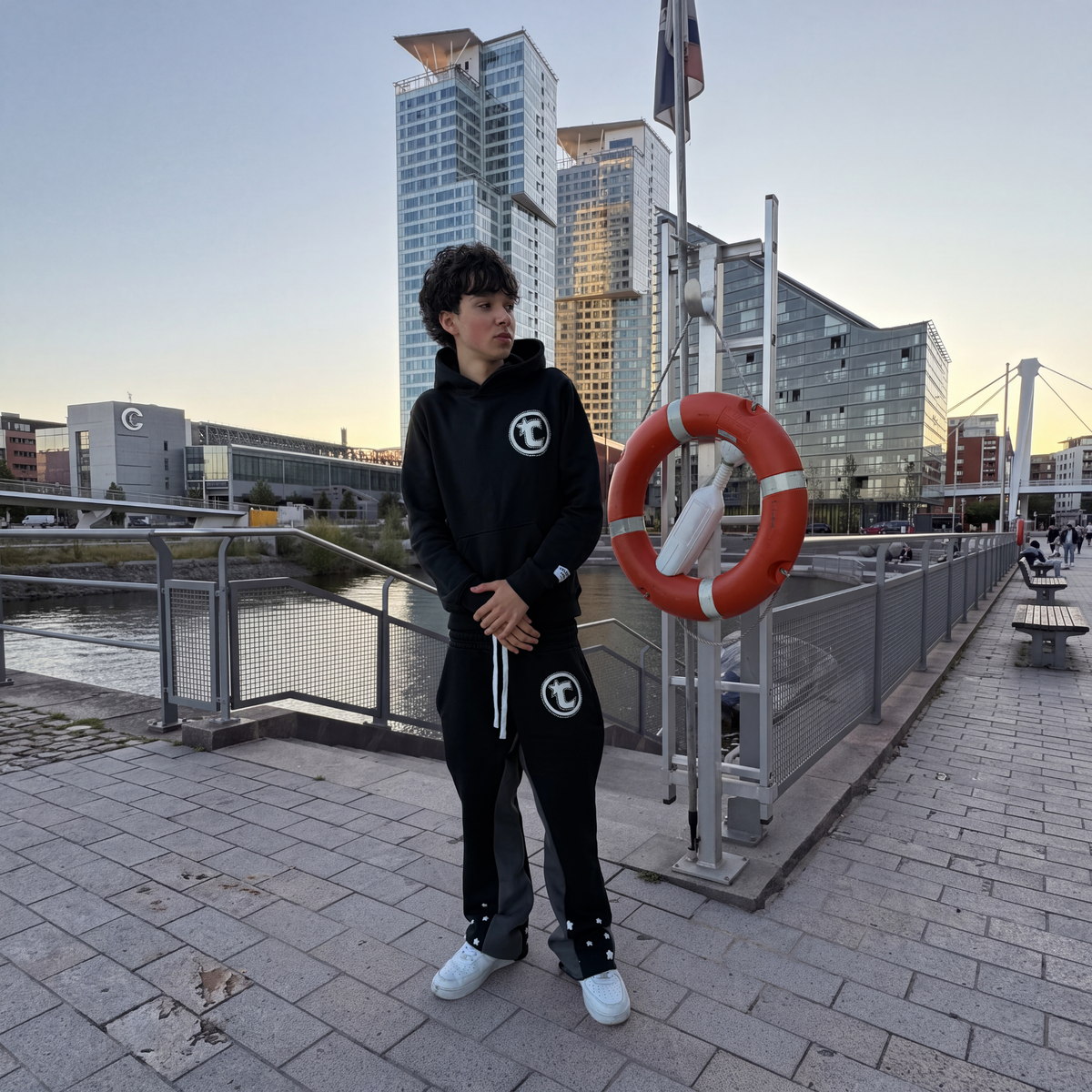 Model wearing the CELESTIC Black Star Hoodie and Black Star Pants by the waterfront, showing the white circular C star logo on the chest and thigh of the heavyweight black set.