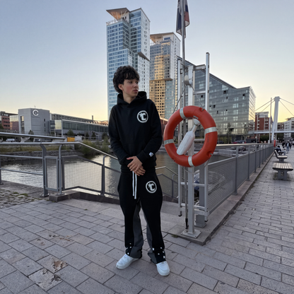 Model wearing the CELESTIC Black Star Hoodie and Black Star Pants by the waterfront, showing the white circular C star logo on the chest and thigh of the heavyweight black set.