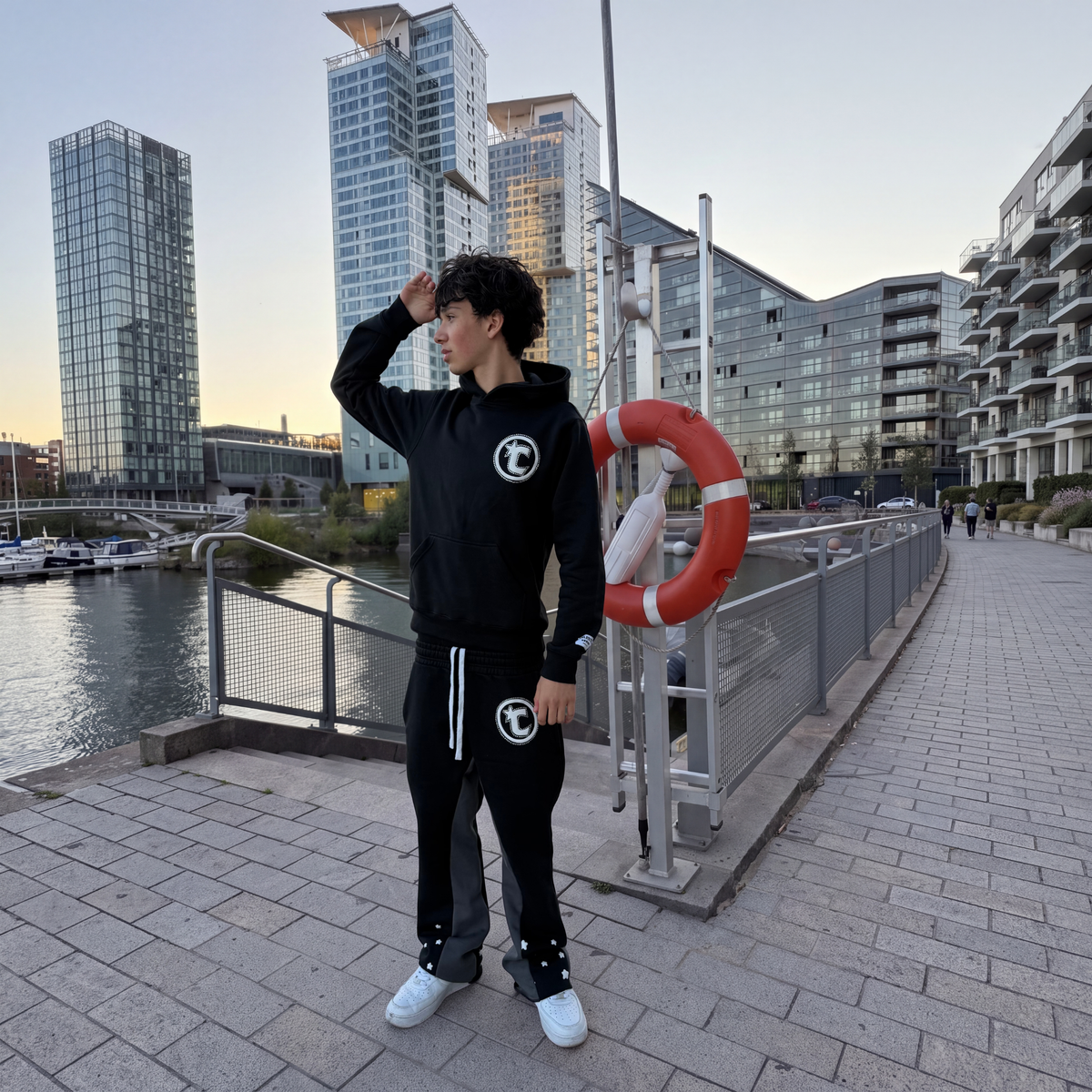 Model posing by the waterfront in the CELESTIC Black Star Hoodie and Black Star Pants, a heavyweight black cotton fleece set with white circular C star logos.