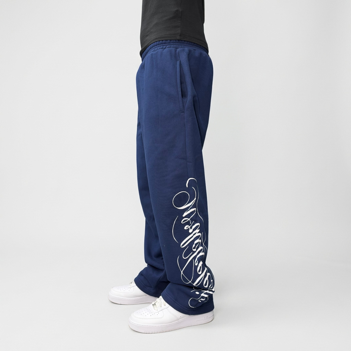 Angled side view on model of the CELESTIC Navy Blue Script Pants with white script print, styled with white sneakers.