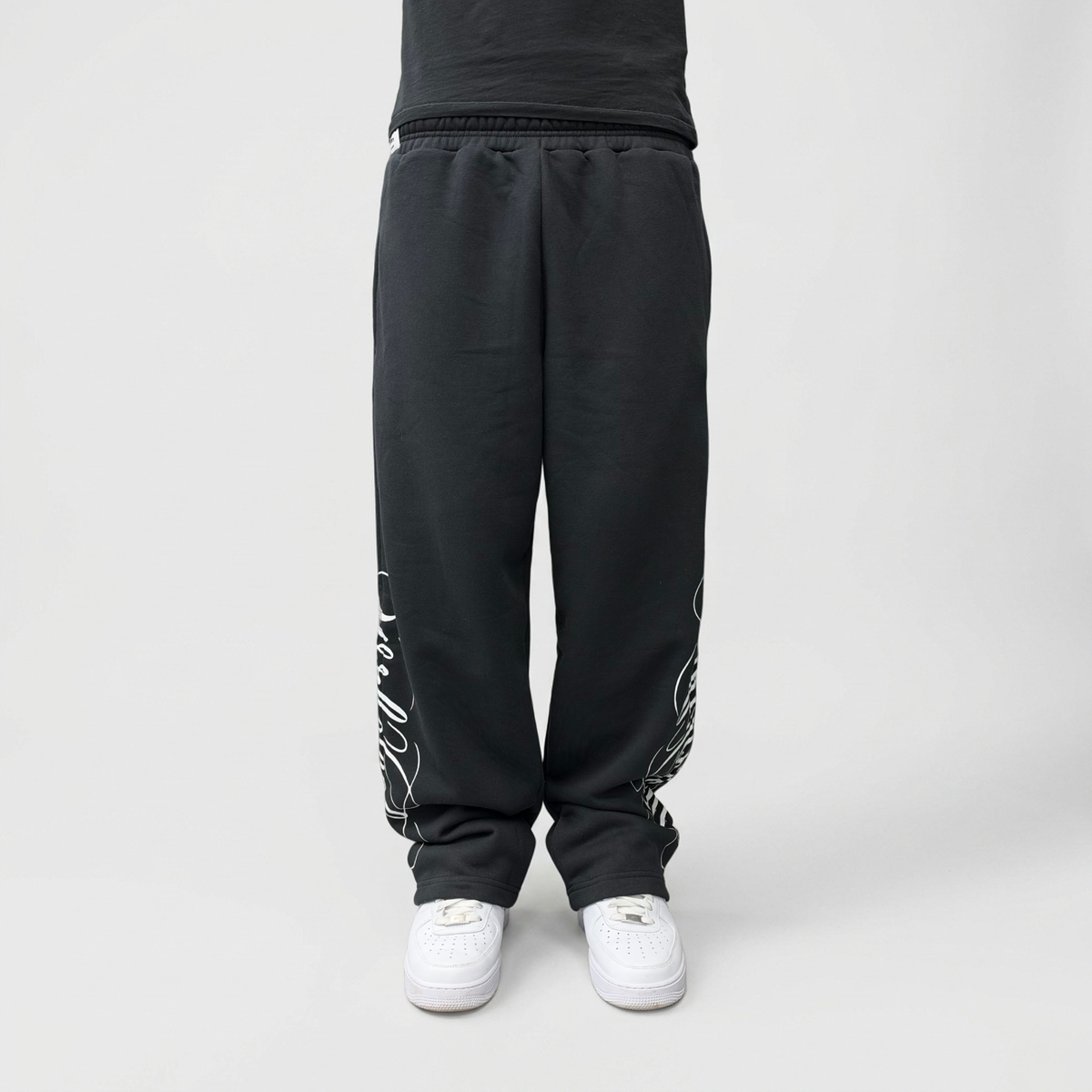 Front view on model of the CELESTIC Black Script Pants in heavyweight black cotton fleece, with white Wrldcelestic script print down the legs.