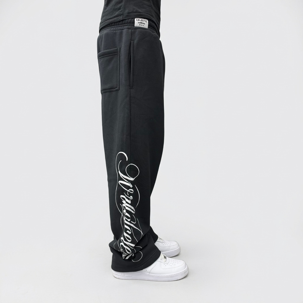 Side view on model of the CELESTIC Black Script Pants, showing the white Wrldcelestic script print running down the leg to the hem.