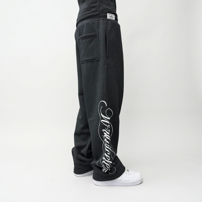 Side view on model of the CELESTIC Black Script Pants in heavyweight black cotton fleece, relaxed straight-leg fit over white sneakers.