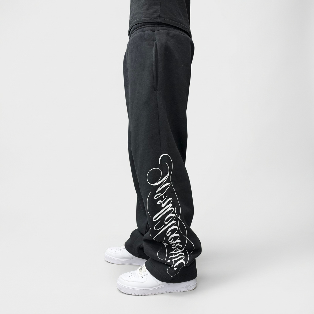 Angled side view on model of the CELESTIC Black Script Pants with white Wrldcelestic script print, styled with white sneakers.