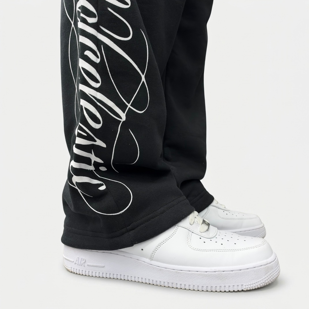 Close-up of the white Wrldcelestic script print on the lower leg of the CELESTIC Black Script Pants, stacking over white sneakers.